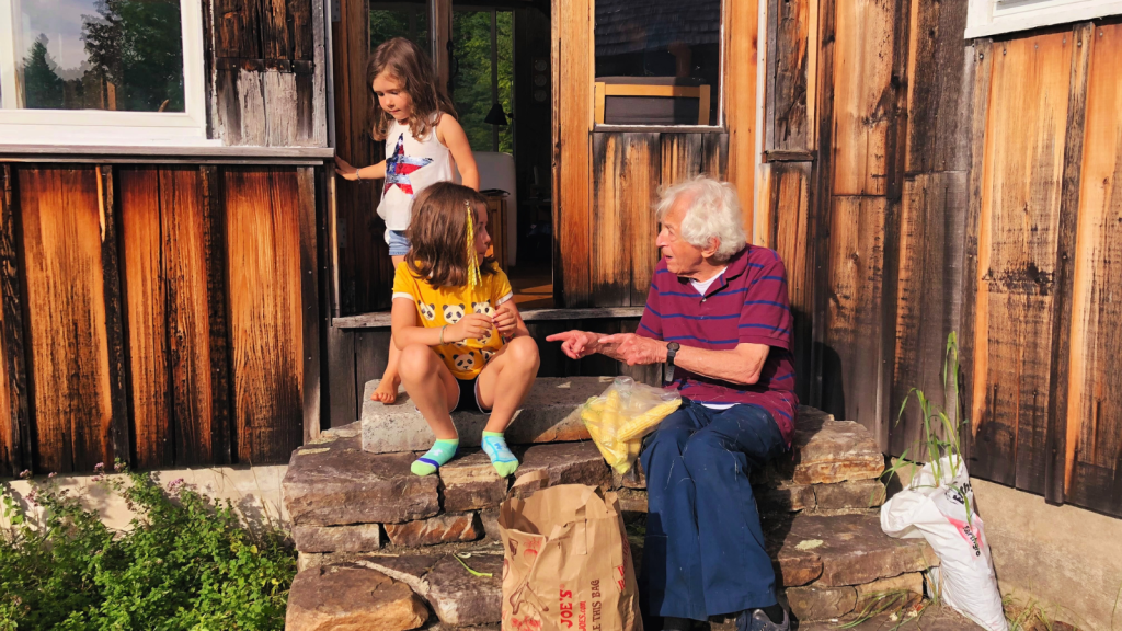 Richard Bernstein, an elderly man in a maroon polo shirt and jeans sits on the back steps of his house shucking corn with two young girls. 